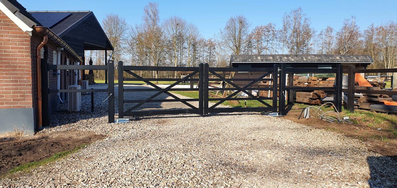 Zwarte stalen landelijke poort met kruisschoren op een erf met grind